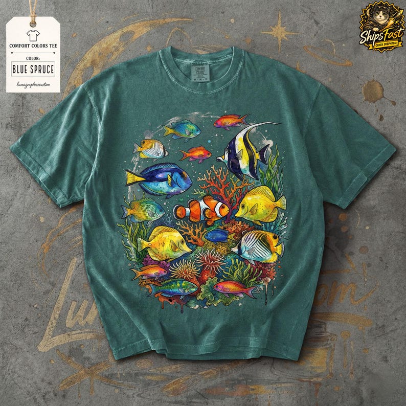 May include: Blue spruce comfort colors tee featuring a colorful illustration of various tropical fish, including clownfish, angelfish, and butterflyfish, swimming around coral. The shirt has a crew neck and short sleeves. The tag reads "Comfort Colors Tee" and "Blue Spruce".