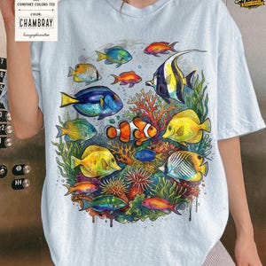 May include: Light blue Comfort Colors tee featuring a vibrant watercolor illustration of various colorful fish and coral. The design includes a clownfish, yellow tang, and other tropical fish. The tag reads "CHAMBRAY" and "lunagraphicscustom".