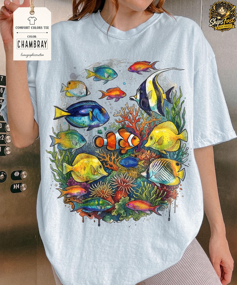 May include: Light blue Comfort Colors tee featuring a vibrant watercolor illustration of various colorful fish and coral. The design includes a clownfish, yellow tang, and other tropical fish. The tag reads "CHAMBRAY" and "lunagraphicscustom".