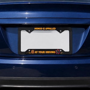 May include: A black license plate frame featuring the yellow text MONGO IS APPALED at the top and AT YOUR DRIVING at the bottom. The frame includes a small graphic of a dog face on the left and a colorful, stylized creature graphic on the right.
