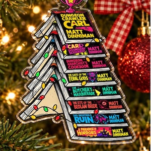May include: A clear acrylic Christmas tree ornament shaped like a stack of Dungeon Crawler Carl books by Matt Dinniman. The design features colourful book spines, a pink trophy topper, and a string of multi-coloured Christmas lights wrapped around.