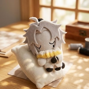 Puede incluir: Una muñeca de peluche con pelo gris y una bufanda amarilla descansa sobre una pequeña almohada blanca. La muñeca tiene los ojos cerrados y pies negros. La almohada está sobre una superficie marrón claro.