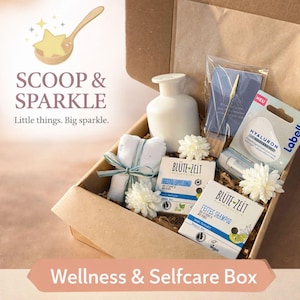 Könnte beinhalten: Eine braune Pappschachtel gefüllt mit Pflegeprodukten. Die Box enthält eine weiße Vase, zwei kleine gerollte Handtücher, die mit einem blauen Band zusammengebunden sind, zwei Seifenstücke und einen Lippenbalsam. Die Box ist mit weißen Blumen und dem Text "SCOOP & SPARKLE" dekoriert.