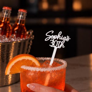May include: A white acrylic drink stirrer featuring the custom text "Sophia's 30th" in a cursive and block font combination, placed inside a glass filled with an orange liquid, ice cubes, and a sugar-rimmed edge, garnished with an orange slice.