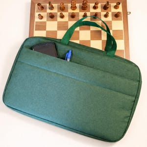 Puede incluir: Una funda de transporte verde de tela para juego de ajedrez con un bolsillo que contiene un teléfono inteligente y un bolígrafo azul. La funda tiene asas verdes y se coloca frente a un tablero de ajedrez de madera con piezas de ajedrez.