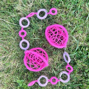 May include: A set of bright pink and lavender crochet jewellery, comprising two necklaces and two spherical ornaments. The necklaces have connected circles and teardrop shapes. The ornaments have a lattice design. The items are displayed on green grass.