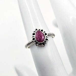 May include: A silver ring featuring a teardrop-shaped magenta stone set in a decorative scalloped metal frame. The band has a textured, twisted rope-like design that encircles the finger, providing a detailed and ornate appearance to the jewelry.