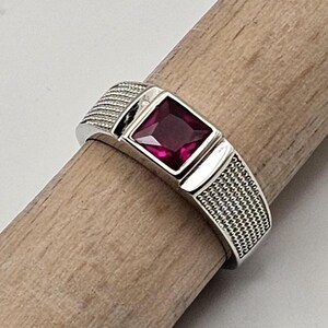 May include: A silver ring with a square, deep red gemstone. The band features a textured design on the sides. The ring is displayed on a wooden dowel against a neutral background. A piece of jewelry.