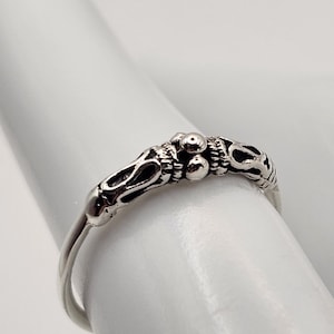 May include: A silver ring with an ornate design. The ring features a detailed pattern of small spheres and curved lines. The ring is displayed on a white surface, showcasing its intricate craftsmanship and elegant style.