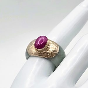 Puede incluir: Un anillo con una banda de jade verde pálido lisa rematada por una placa de metal dorado texturizado y martillado. Centrada en la placa hay una piedra cabujón ovalada de color magenta intenso engastada en un delicado borde de metal dorado.