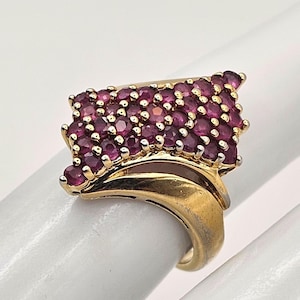May include: A gold-toned ring featuring a cluster of small, round, deep red gemstones. The gemstones are set in a rectangular shape, with a curved band. The ring is displayed on a white surface.
