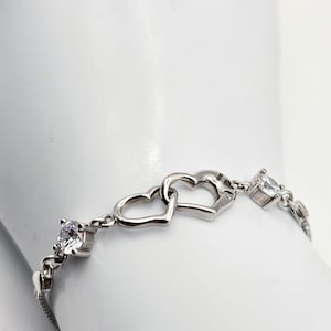May include: A silver-toned bracelet featuring two interlocking heart-shaped links at the center, flanked by clear, round-cut stones set in metal prongs. The chain consists of delicate metal links that connect the central hearts and the stones.