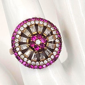 May include: A circular ring featuring a central cluster of round pink stones and one white stone, surrounded by a ring of baguette-cut white stones, a ring of round white stones, and an outer border of round pink stones set in a gold-tone metal.
