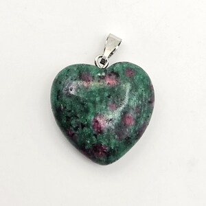 May include: A heart-shaped pendant made of polished zoisite stone with deep green and magenta-colored ruby inclusions. The stone is smooth and rounded, featuring a silver-toned metal bail attached to the top for hanging on a chain or cord.