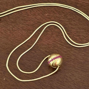 May include: A gold-toned snake chain necklace featuring a spherical pendant. The pendant is smooth and metallic with a horizontal band of small, rectangular, vibrant pink stones encircling the center of the sphere. The chain has a lobster clasp.