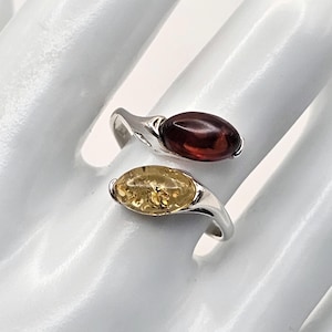 May include: A silver bypass ring featuring two marquise-cut stones. One stone is a deep, translucent dark red amber, and the other is a lighter, translucent golden yellow amber. The band is polished silver, creating a sleek and continuous design.