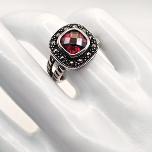 May include: A silver ring featuring a square-cut, faceted deep red gemstone at the center. The stone is surrounded by a border of small, dark, metallic stones. The band has a textured, ribbed design on the sides, creating a detailed appearance.
