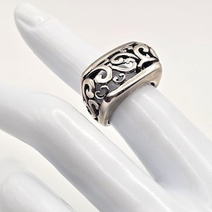 May include: A wide silver ring featuring an intricate openwork filigree design with swirling patterns. The band has a polished finish and a substantial width, showcasing a detailed cutout metalwork motif that wraps around the entire circumference.