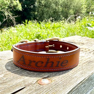 May include: A brown leather dog collar with the name "Archie" engraved on the front. The collar has brass-coloured hardware and blue stitching. The collar is on a weathered wooden surface.