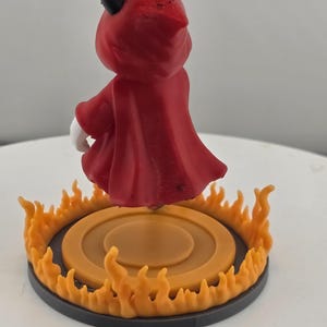 May include: A small figurine of a character wearing a red hooded cloak with black horns, standing on a circular base surrounded by orange flame-like accents. The character has white hands and is positioned facing away from the viewer.