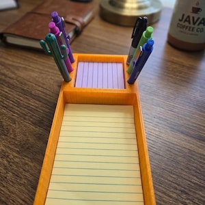 May include: An orange desk organizer with a rectangular base holding a large yellow lined notepad and a smaller square purple notepad. The organizer features integrated slots for writing instruments, including a green cactus-shaped pen.