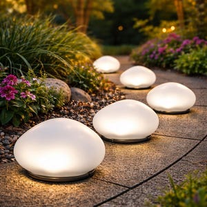 Puede incluir: Camino de jardín iluminado por luces exteriores blancas en forma de guijarro. Las luces están espaciadas a lo largo de un sendero de piedra, rodeado de vegetación y flores. Luces de cadena crean un ambiente cálido.