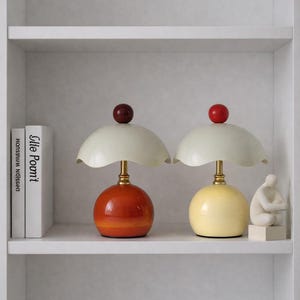 以下が含まれることがあります： レトロなデザインの装飾的なテーブルランプ2つ。1つのランプはオレンジ色のベース、もう1つは黄色のベースです。どちらのランプも白いランプシェードと、上部に赤または栗色の球体が付いています。ランプは白い棚に置かれています。