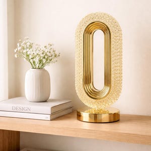 Puede incluir: Lámpara de mesa dorada de forma ovalada con una superficie texturizada y cristalina. La lámpara tiene una base circular y una serie de marcos ovalados concéntricos en su interior. También se ve un jarrón blanco con flores y libros con la etiqueta "DESIGN".