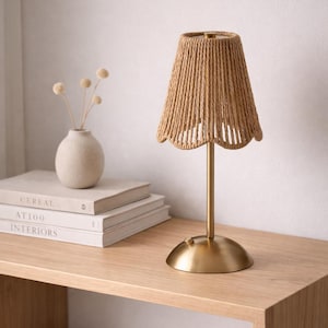 Może przedstawiać: Lampa stołowa z tkanym abażurem w naturalnym beżowym kolorze. Lampa ma złotą podstawę i trzon. Stoi na drewnianym stole obok książek i małego wazonu z suszonymi kwiatami. Wysokość lampy to około 30 cm.