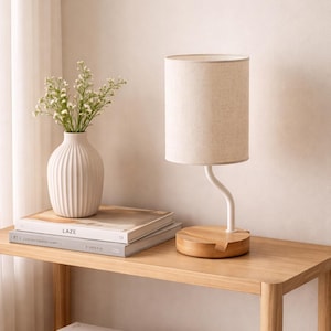 Op de afbeelding: Een moderne tafellamp met een beige stoffen kap en een witte, gebogen steel staat op een houten voet. Een witte vaas met bloemen en boeken staan op een lichte houten tafel. Het woord "LAZE" is zichtbaar op een van de boeken.