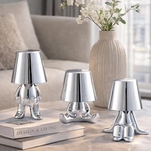 Può includere: Tre lampade da tavolo cromate con un design umanoide unico. Ogni lampada presenta un paralume conico sopra una figura stilizzata con braccia e gambe. Le lampade sono disposte su un tavolo con un vaso di fiori bianchi e un libro "DESIGN & LIVING".