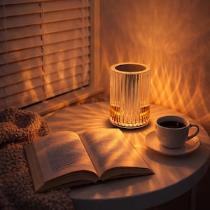 May include: A cylindrical, ribbed glass lamp emits a warm light, illuminating an open book and a cup of coffee on a white table. A cosy blanket and closed blinds are also visible. The lamp is a decorative item.