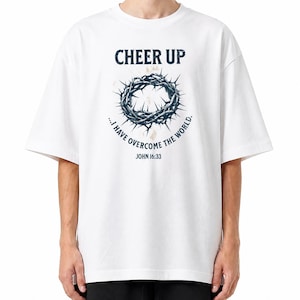 Op de afbeelding: Een wit t-shirt met korte mouwen en een donkerblauwe afbeelding van een doornenkroon. De tekst luidt CHEER UP, ...I HAVE OVERCOME THE WORLD en JOHN 16:33. Het ontwerp is gecentreerd op de borst met een verweerde textuur op de illustratie.