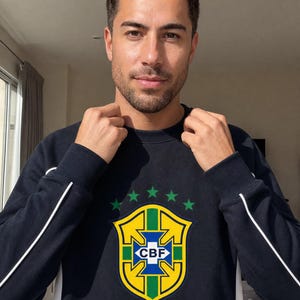 Könnte beinhalten: Ein dunkelblaues Langarm-Sweatshirt mit einem weißen vertikalen Streifen an den Seiten und dem CBF Brasil-Logo auf der Brust. Das Logo besteht aus einem gelben Schild mit grünen Akzenten, fünf grünen Sternen darüber und dem Text CBF und BRASIL.