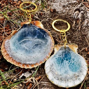 May include: Two seashell keychains with gold-colored hardware. The shells have a resin interior with blue and white swirls, resembling ocean waves. The edges of the shells are a natural brown color. The keychains are displayed on a natural outdoor surface.