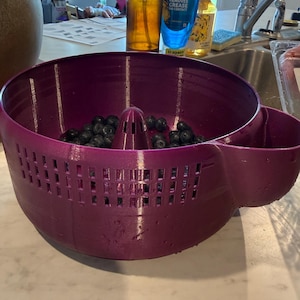 May include: A deep purple berry washing bowl with a built-in colander and side container. The bowl is filled with fresh blueberries. The bowl has a perforated design for draining water.