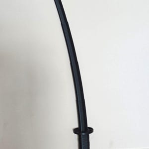 May include: A black katana sword with a curved blade. The handle is textured, and the guard is a circular design. The sword is leaning against a white wall.