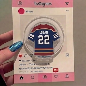 May include: A clear, circular acrylic magnet featuring a navy blue and maroon jersey design with the name "LOGAN" and the number "22". The magnet is set against a pink Instagram-themed background with text and icons.