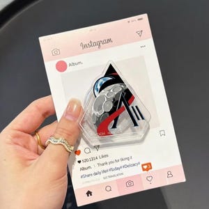 May include: A clear acrylic phone grip with a design featuring a moon, a planet, and the letter "A". The grip is held against a pink Instagram-themed background. The grip has the words "WISEMAN", "OLOVER", and "KOCH HANSEN" printed on it.