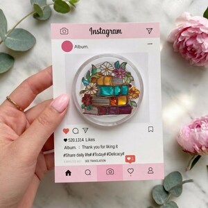 May include: A circular phone grip featuring a stack of colorful books adorned with flowers, mounted on a rectangular card designed like an Instagram post. The card displays the text Album, 520,1314 Likes, and hashtags #Share daily life #Today #Delicacy.