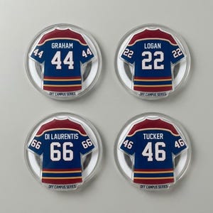 May include: Four circular magnets featuring miniature hockey jerseys. Each jersey is navy blue with white numbers and a red and gold trim. The names Graham, Logan, Di Laurentis, and Tucker are printed above the numbers. The text "OFF CAMPUS SERIES" is printed at the bottom of each magnet.