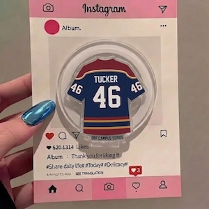 May include: A clear acrylic phone grip in the shape of a blue and red jersey, featuring the name "TUCKER" and the number "46". The grip is set against a pink Instagram-themed background with text including "Album" and "520.1314 Likes".