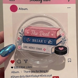 May include: A clear circular phone grip featuring a stack of three books with pink, blue, and white covers. The books are labeled The Deal, BRIAR U, and LOVE. HOCKEY. CHAOS. A black hockey puck with a pink heart is positioned next to the books.