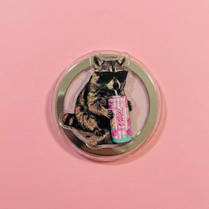 May include: A round phone grip featuring a raccoon wearing sunglasses and drinking from a can. The can is pink, blue, and white with the word "Aloha" printed on it. The background is pink.