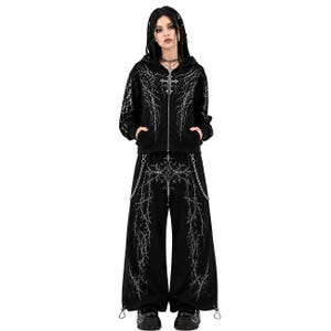 Puede incluir: Un conjunto de sudadera negra con cremallera y pantalones anchos a juego, con un estampado gráfico plateado que recuerda a rayos. La sudadera incluye cremallera frontal y capucha, mientras que los pantalones tienen cadenas plateadas y cordones.