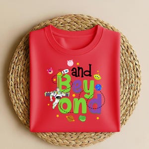 Może przedstawiać: Czerwona koszulka z napisem "and Beyond" w zabawnej czcionce z kresk&oacute;wkową grafiką rakiety, kosmity i innych element&oacute;w o tematyce kosmicznej. Koszulka jest złożona i umieszczona na plecionej macie.