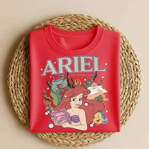 Puede incluir: Una sudadera roja con un gr&aacute;fico de Ariel de La Sirenita. El dise&ntilde;o incluye a Ariel, Flounder, el Rey Trit&oacute;n y las palabras "ARIEL THE PRINCESS" en un estilo vintage. La sudadera se muestra sobre una estera tejida.