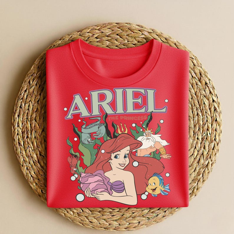 Puede incluir: Una sudadera roja con un gr&aacute;fico de Ariel de La Sirenita. El dise&ntilde;o incluye a Ariel, Flounder, el Rey Trit&oacute;n y las palabras "ARIEL THE PRINCESS" en un estilo vintage. La sudadera se muestra sobre una estera tejida.