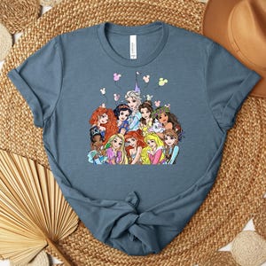 May include: A blue t-shirt featuring a colorful graphic of various cartoon princesses. The princesses are depicted with detailed facial features and vibrant dresses. The shirt has a classic crew neck and short sleeves, suitable for casual wear.