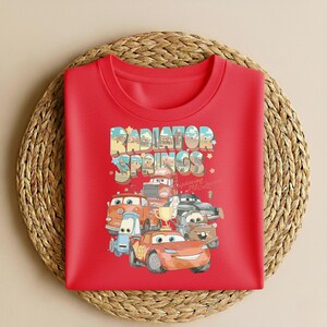 May include: A red crew neck sweatshirt featuring a colorful graphic of characters from the movie Cars. The graphic includes the text "Radiator Springs" in a stylized font, along with images of various cars. The sweatshirt is displayed on a woven surface.